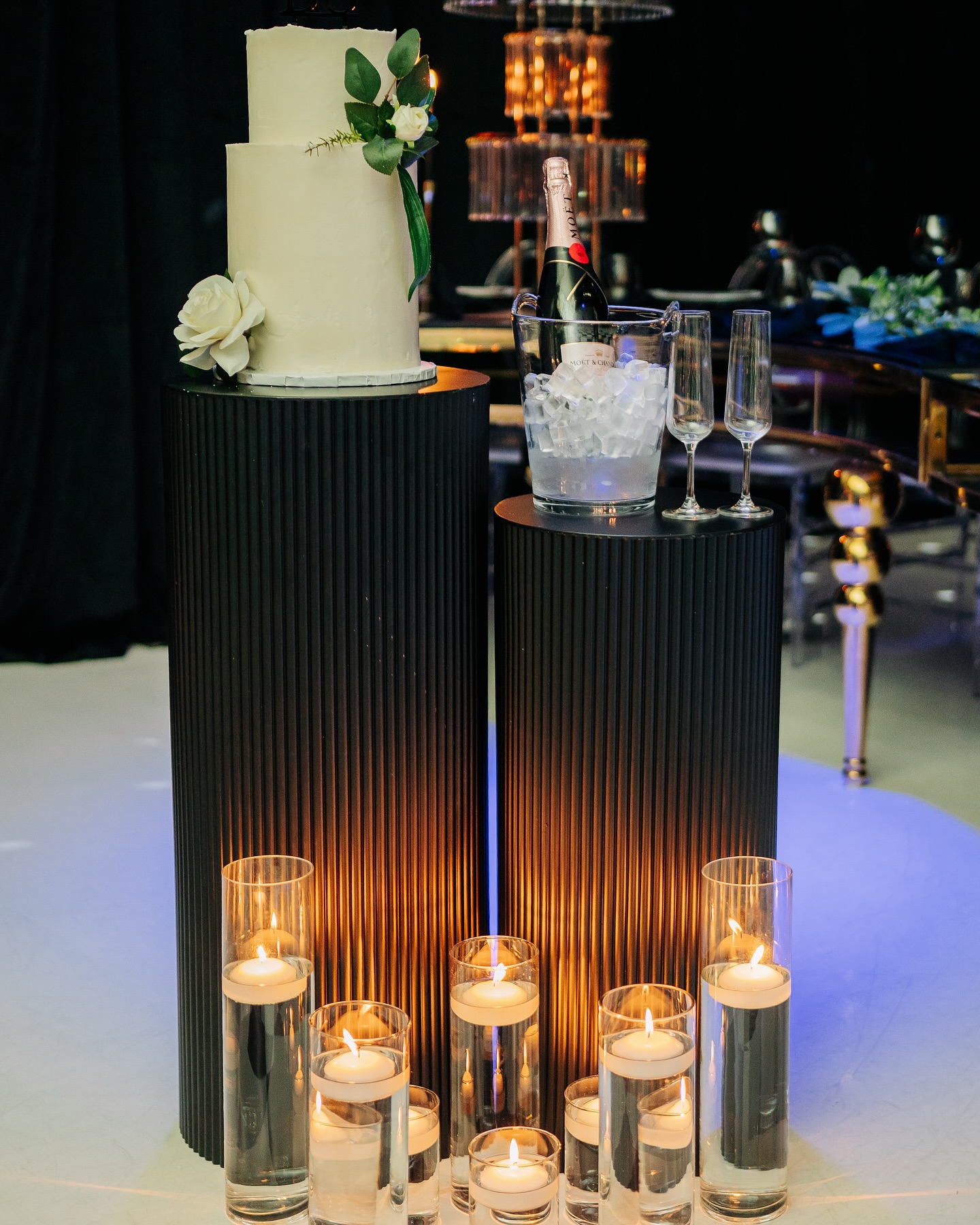 Tiered white celebration cake and champagne display arranged on fluted white pedestals with flowers and candles.